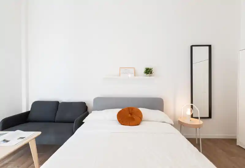 Camera da letto luminosa e minimale con letto matrimoniale centrale, cuscino rotondo decorativo, comodino con lampada e piccolo divano a sinistra — perfetta per un annuncio moderno e pulito.