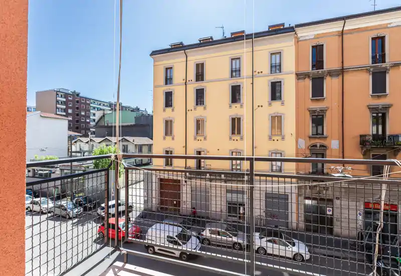 Balcone urbano con vista sulla strada e sugli edifici residenziali vicini; ringhiera metallica e pannelli in vetro offrono uno spazio esterno luminoso e sicuro.