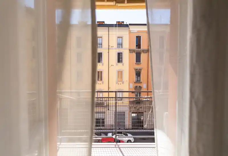 Piccolo balcone incorniciato da tende leggere, con vista su una strada e facciate di palazzi storici e auto parcheggiate; la luce naturale morbida crea un’atmosfera urbana accogliente.