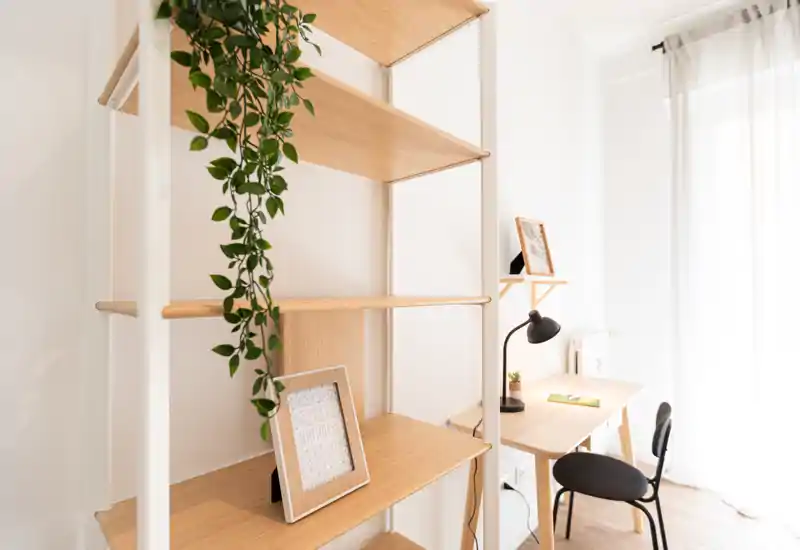 Zona giorno luminosa e minimal con una libreria in legno e una piccola scrivania vicino a una finestra soleggiata — ideale come angolo studio o lettura.