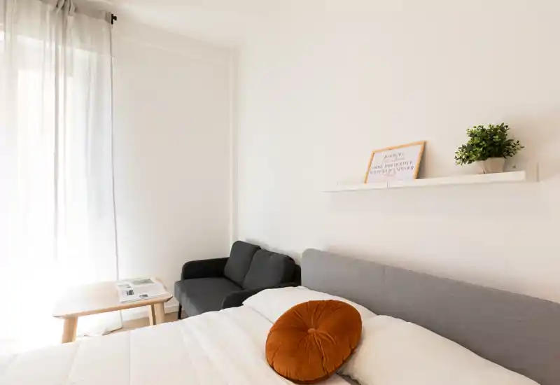 Camera da letto luminosa e minimal con letto ben rifatto, cuscino decorativo rotondo e un piccolo divano accanto alla finestra che lascia entrare molta luce naturale.