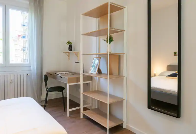 Rincón de dormitorio luminoso y moderno con vista parcial de la cama, escritorio compacto junto a la ventana y estantería abierta. Decoración limpia y minimalista, luz natural y detalles cálidos, ideal para un anuncio de alquiler.