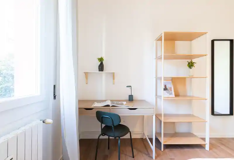 Rincón de trabajo luminoso y minimalista con escritorio de madera, silla y estantería junto a una gran ventana que aporta luz natural.