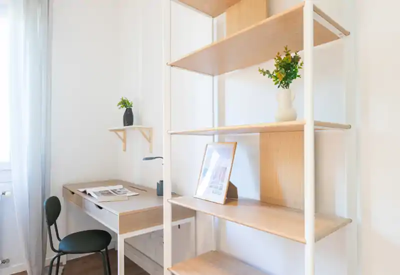 Rincón de estudio luminoso y minimalista con escritorio de madera, silla y estantería abierta — ideal para teletrabajo o lectura, realzado por la luz natural y detalles decorativos sencillos.