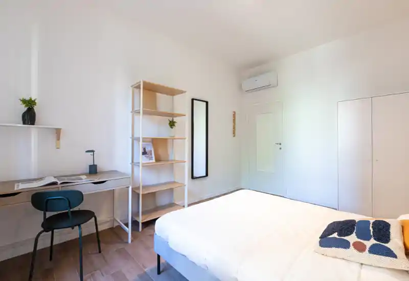Dormitorio luminoso y minimalista con cama doble bien hecha, suelo de madera, estantería y un pequeño escritorio — ideal para un viajero solo o pareja que busque un espacio moderno y tranquilo.
