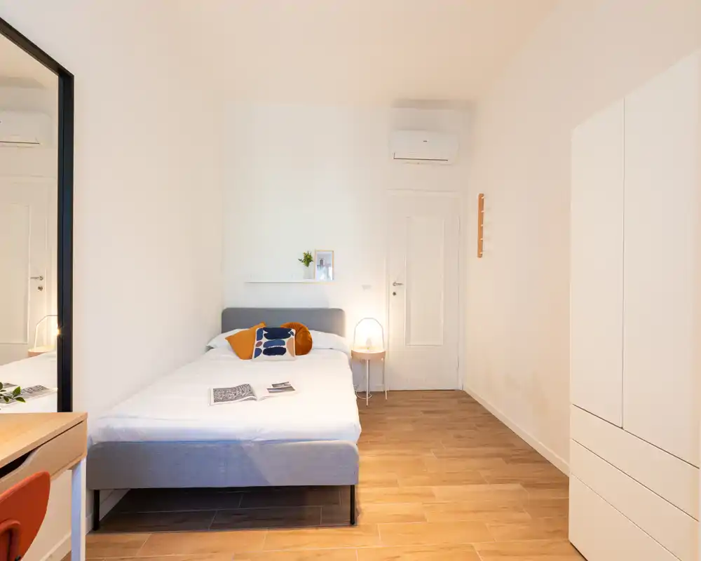 Camera da letto minimalista e luminosa con letto matrimoniale, lampada da comodino, mensola semplice e armadio — pavimento in legno caldo e arredo pulito creano un'atmosfera accogliente.