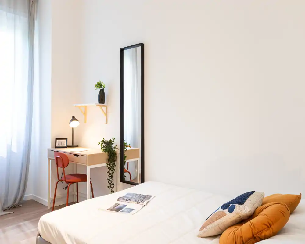 Camera da letto luminosa e minimal con un letto matrimoniale ben rifatto e cuscini decorativi, una piccola scrivania vicino alla finestra e uno specchio alto che amplia la stanza.