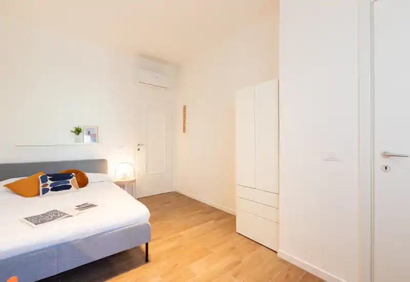 Camera da letto moderna e luminosa con letto matrimoniale ben rifatto, cuscini decorativi caldi e lampada da comodino. Armadio semplice e pavimento in legno creano un’atmosfera pulita e accogliente, perfetta per affitti brevi.