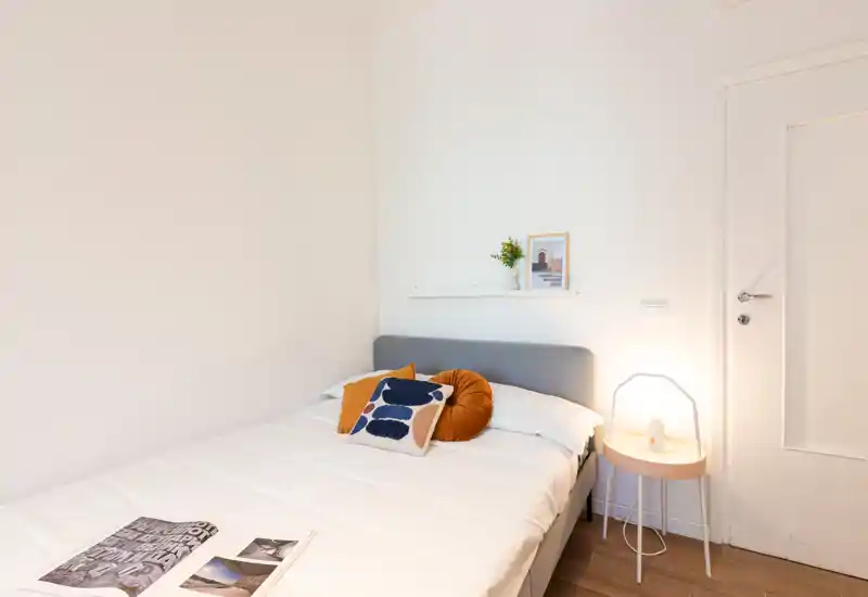 Camera da letto luminosa e minimal con letto matrimoniale confortevole, cuscini decorativi e una lampada da comodino calda — perfetta per un soggiorno accogliente e moderno.