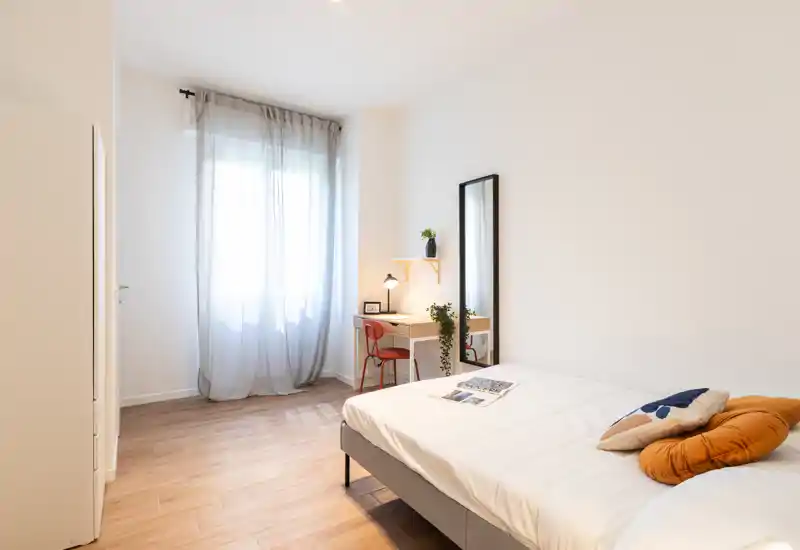 Camera da letto luminosa e minimale con un comodo letto matrimoniale, tessuti morbidi e una piccola scrivania vicino alla finestra. La luce naturale attraverso le tende leggere e il pavimento in legno creano un'atmosfera accogliente.