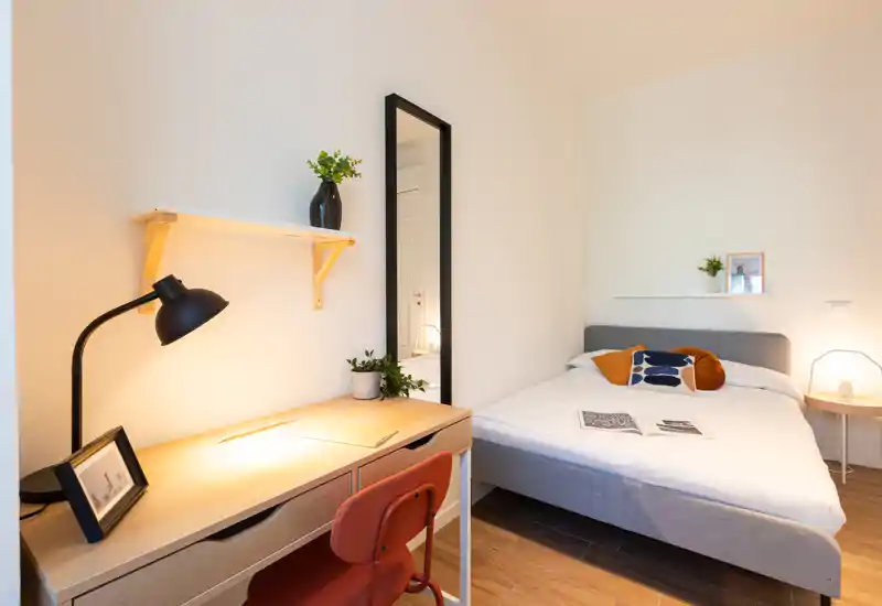 Camera da letto moderna e luminosa con letto matrimoniale rifatto, cuscini decorativi, lampada da comodino e una scrivania in legno con lampada: ideale per riposo e lavoro.