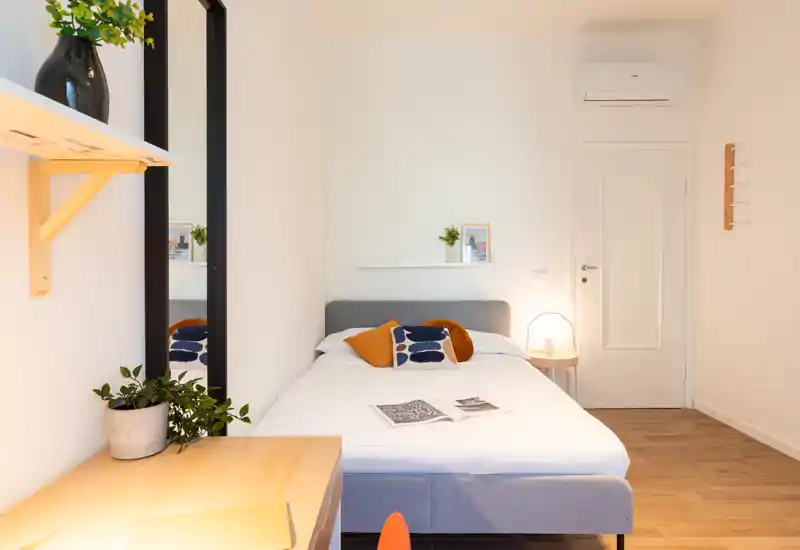 Camera da letto luminosa e ben allestita con letto matrimoniale ordinato, cuscini decorativi, lampada da comodino e pavimento in legno caldo — ideale per annunci.