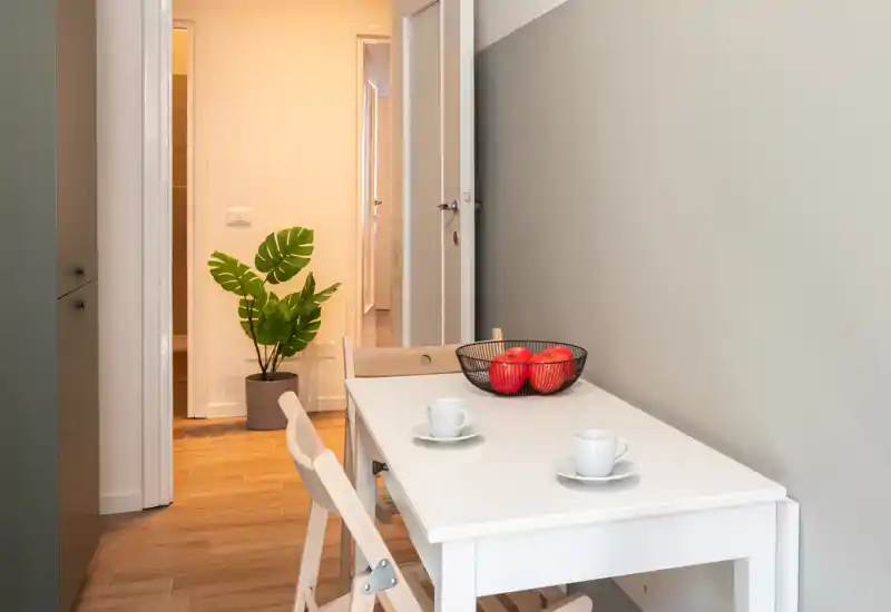 Angolo pranzo compatto con tavolo bianco per due, ciotola di frutta decorativa e pianta in vaso; pavimento in legno caldo e pareti neutre per un’atmosfera moderna e tranquilla.