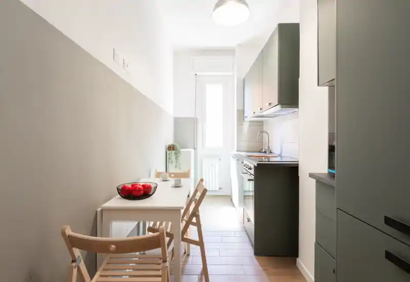 Cucina moderna e luminosa, stretta ma funzionale, con pensili integrati, un piccolo tavolo da pranzo e sedie pieghevoli in legno; la luce naturale dalla finestra la rende ariosa.