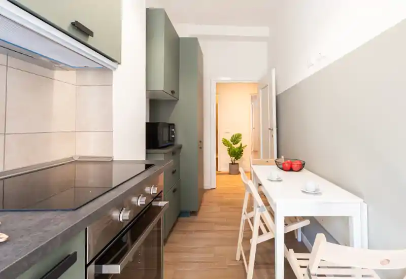 Cucina moderna a corridoio con pensili verde oliva, piano a induzione e piccolo tavolo da pranzo per due; ambiente luminoso e ben allestito, ideale per appartamenti compatti.