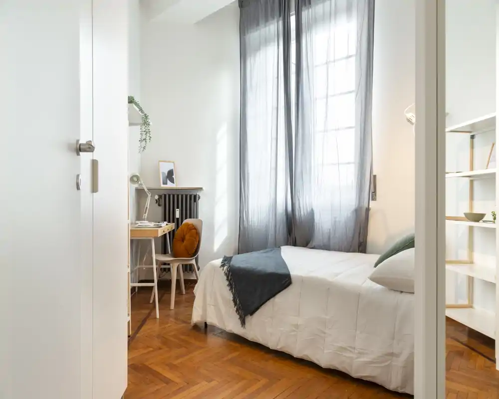 Bright, stylish bedroom with a neatly made bed, large window with sheer curtains, and a cozy reading desk — ideal for rental photos.