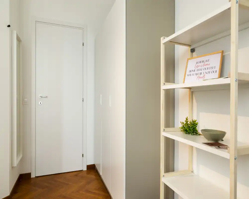 Minimal entrance/hallway with a white door, built-in wardrobe and a decorative open shelf; warm wood parquet flooring and soft neutral tones create a clean, welcoming first impression.
