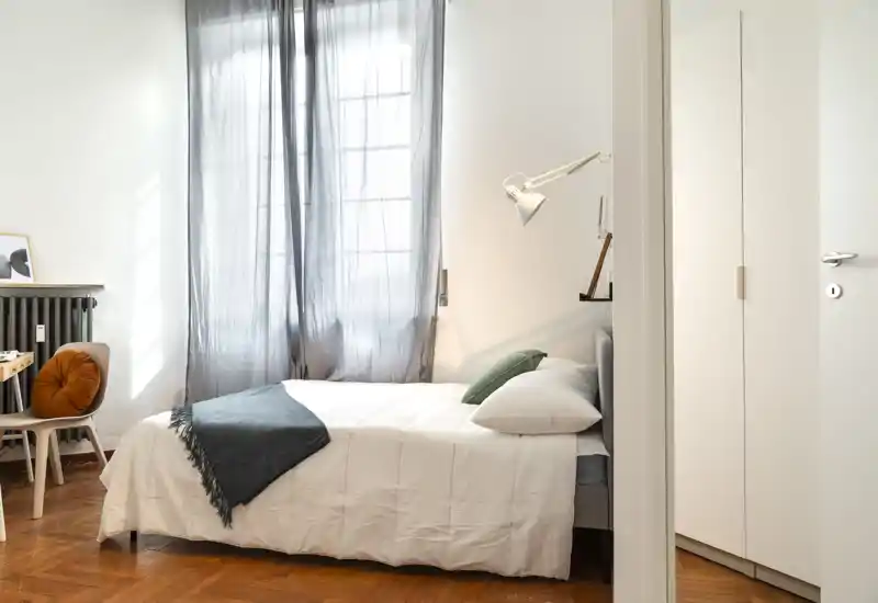 Bright, minimalist bedroom with a neatly made double bed, soft white linens, a green accent pillow and a grey throw by a large window with sheer curtains.