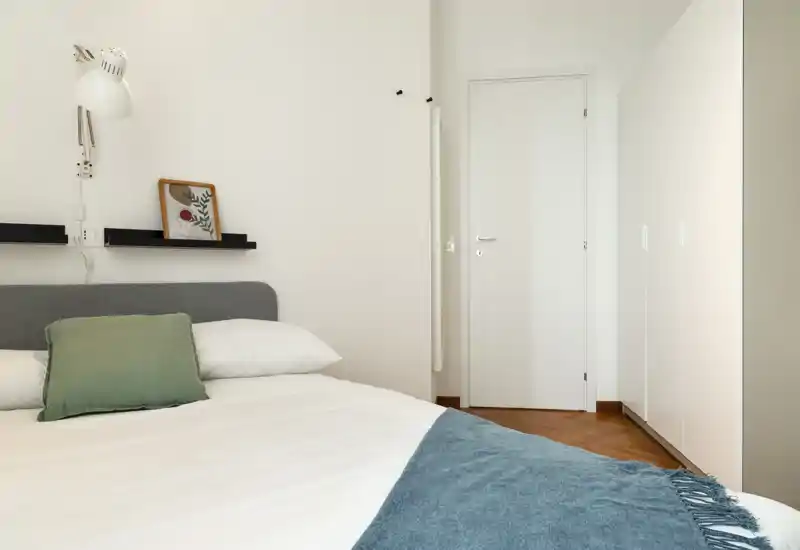 Bright, modern bedroom with a neatly made bed, decorative cushion and blue throw; simple white walls and a wardrobe create a clean, minimal look.