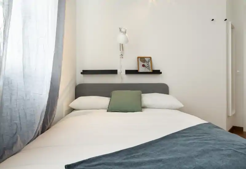 Bright, minimal bedroom with a neatly made double bed, cozy throw and soft natural light from a window to the left.