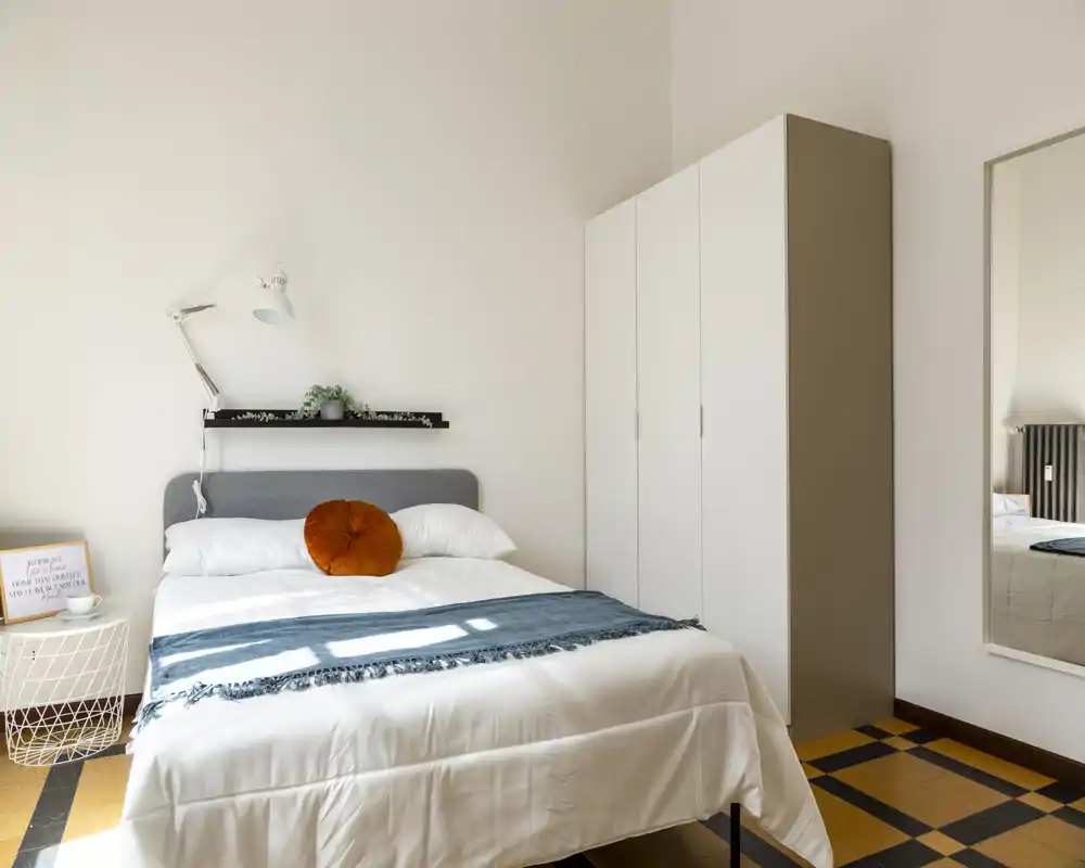 Bright, modern bedroom with a neatly made double bed, decorative round cushion and a wardrobe. Natural light fills the room, creating a calm and welcoming atmosphere.
