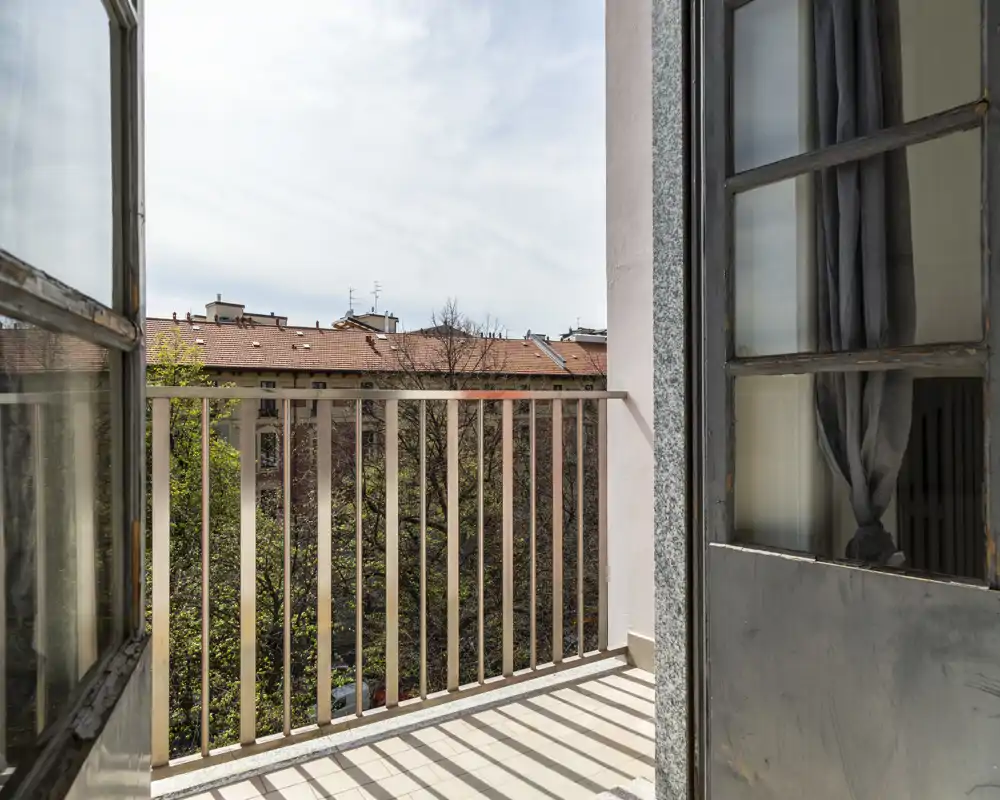 Sunny balcony with metal railing overlooking tree tops and neighboring rooftops; tiled floor and open double doors bring in natural light—ideal to show outdoor space.