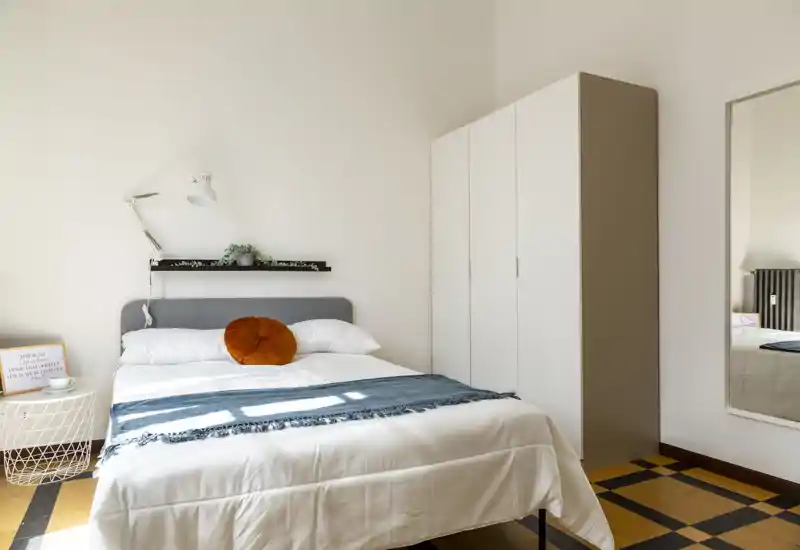 Bright, modern bedroom with a neatly made double bed, decorative round cushion and a wardrobe. Natural light fills the room, creating a calm and welcoming atmosphere.