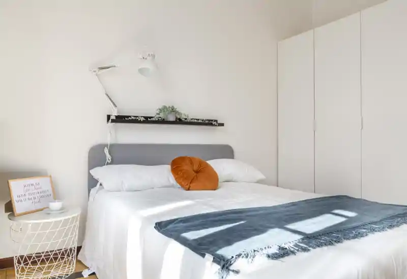Bright, modern bedroom with a fully visible double bed, soft gray headboard, decorative orange cushion and a blue throw; built-in wardrobe and minimalist shelving complete the airy layout.
