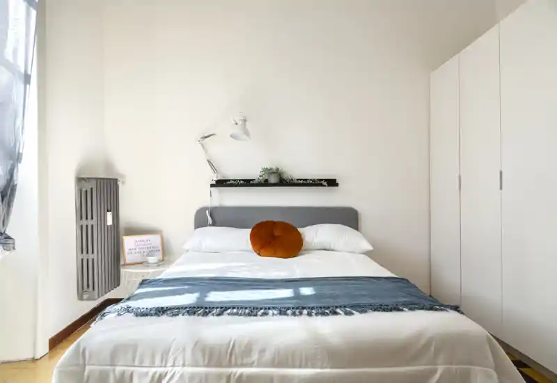 Bright, minimalist bedroom with a neatly made double bed, a blue throw and a decorative round cushion; wardrobe to the right and natural light flooding the room.