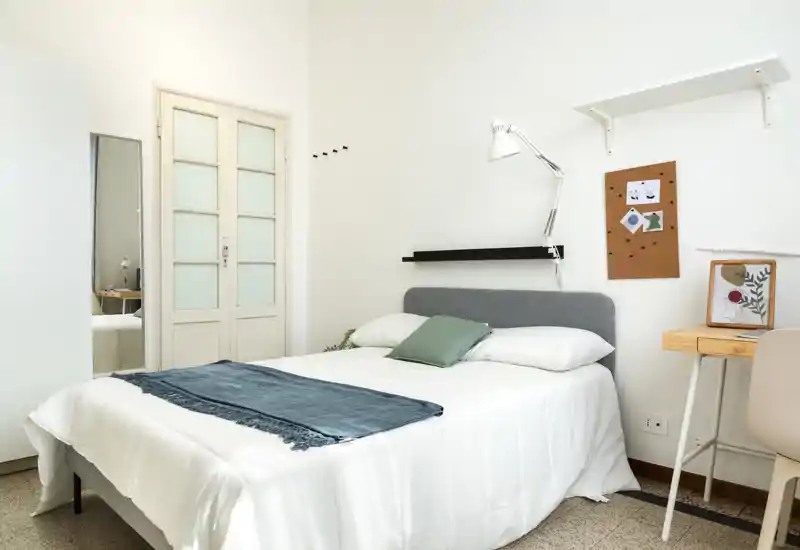 Bright, minimal bedroom with a neatly made double bed, soft linens and a small study desk — ideal for a comfortable stay.
