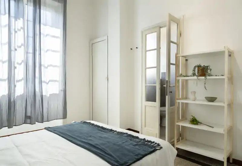 Bright, minimalist bedroom with a neatly made bed, soft throw and natural light from a large window; simple shelving and neutral decor create a calm atmosphere.