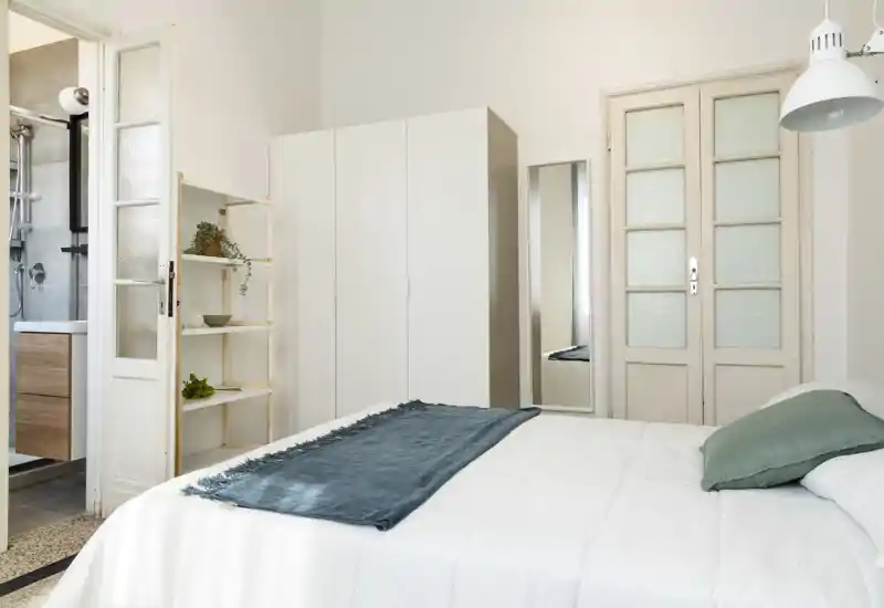 Bright, minimalist bedroom with a neatly made bed, wardrobe, full-length mirror and a door leading to an ensuite bathroom. Neutral tones and simple decor create a clean, airy atmosphere ideal for rental listings.