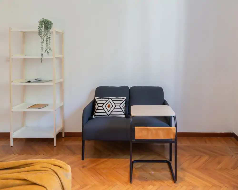 Zona de estar compacta con sofá de dos plazas, mesa auxiliar y estantería abierta; suelo de parqué cálido y luz natural suave que crean un ambiente acogedor.
