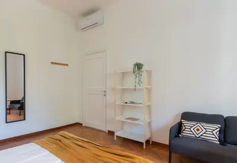 Dormitorio luminoso y minimalista con cama bien hecha, una manta cálida y una pequeña estantería. Un sillón y un espejo completan este espacio acogedor y bien iluminado.