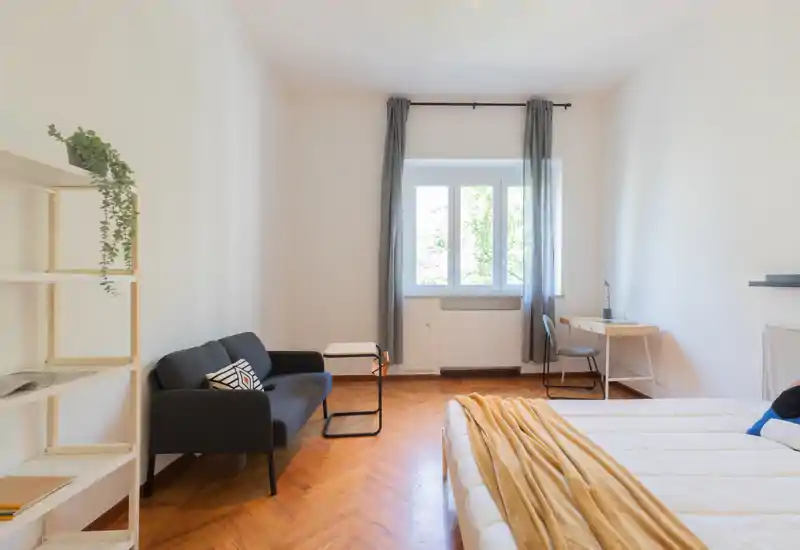 Dormitorio luminoso y moderno con una cama cómoda, un pequeño sofá y un escritorio junto a la ventana — ideal para alquileres temporales. Suelo de parquet en espiga y decoración neutra crean un ambiente tranquilo y acogedor.