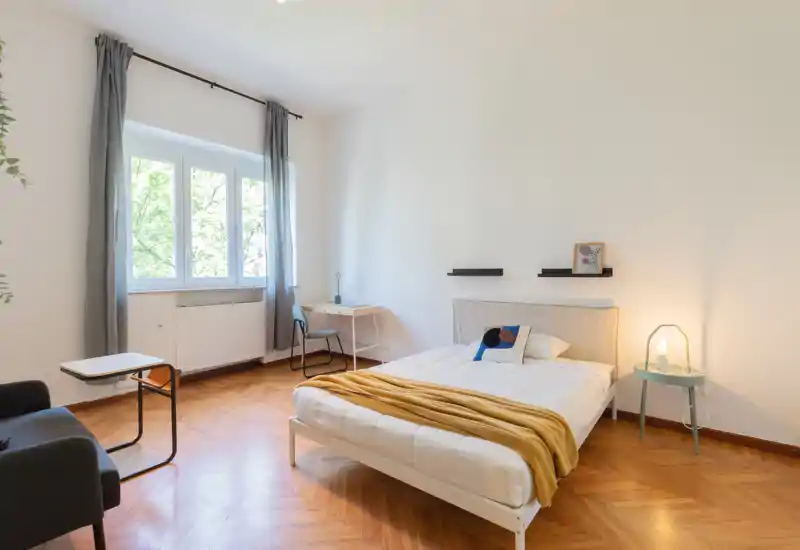 Dormitorio luminoso y minimalista con cama doble, suelo de parquet cálido, mesita y escritorio junto a la ventana. Decorado con gusto con una manta mostaza y luz cálida que lo hace acogedor.