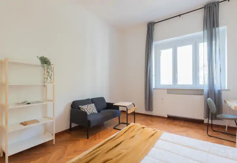Dormitorio luminoso y minimalista con cama en primer plano, un pequeño sofá y mesa auxiliar junto a la pared y una gran ventana que proporciona luz natural.