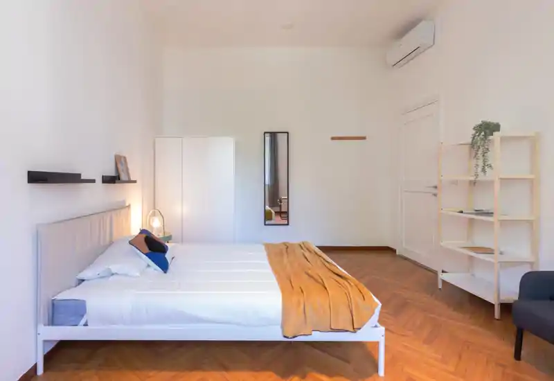 Dormitorio luminoso y minimalista con cama doble totalmente visible, suelo de parquet cálido y textiles cuidados como una manta mostaza y cojines; incluye armario, espejo y estantería — perfecto para anuncios que destaquen limpieza y comodidad moderna.