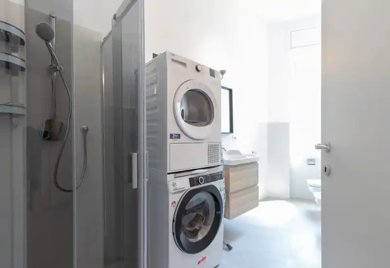 Bright, modern bathroom with a stacked washer and dryer, glass shower enclosure, wall-mounted sink and natural light from a high window.