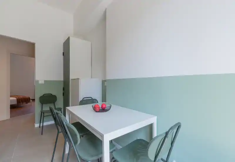 Bright, modern dining corner with a white table, four green metal chairs and a simple fruit bowl; soft two-tone wall paint adds a calm, contemporary feel.