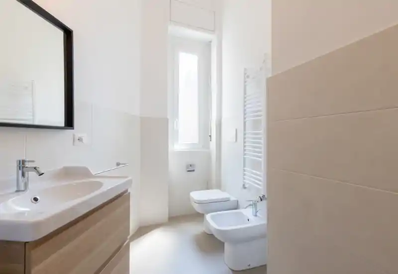 Bright, modern bathroom with a sink and vanity, bidet and toilet beside a tall frosted window; clean tiles and a wall-mounted towel warmer create a fresh, airy feel.
