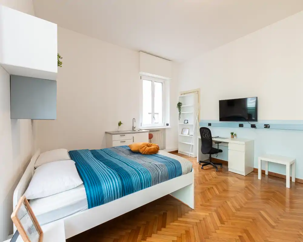 Camera da letto luminosa e moderna con letto matrimoniale, pavimento in parquet e scrivania con TV a parete — ideale per soggiorni comodi o studenti.