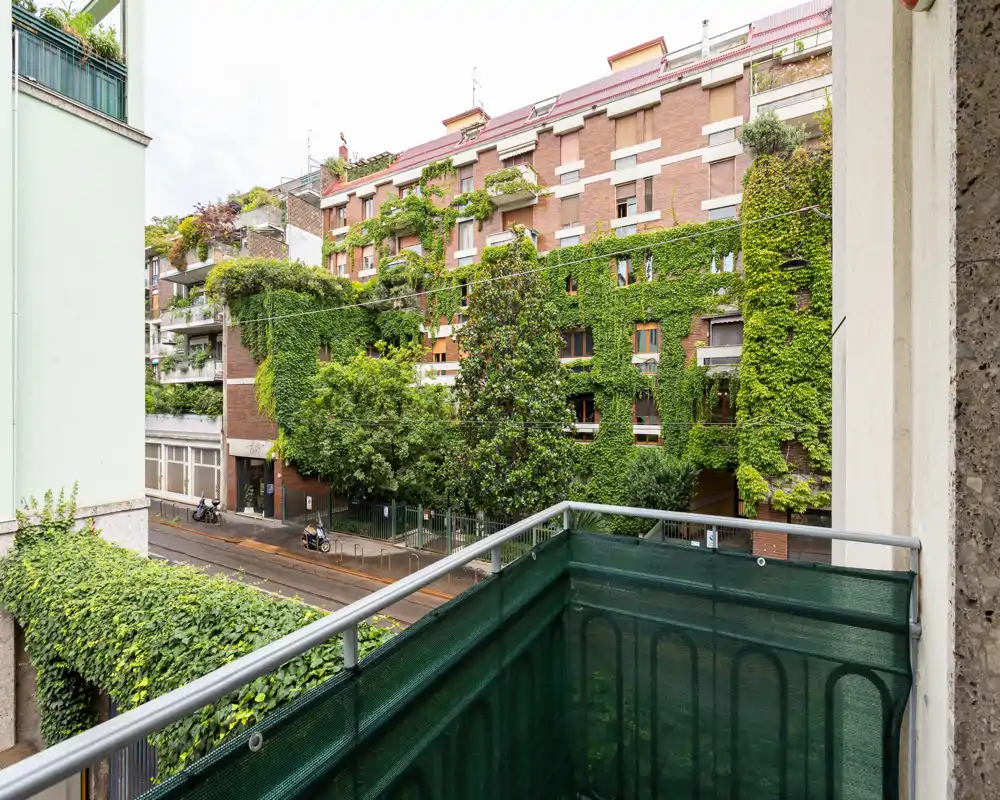 Piccolo balcone con vista su una strada alberata e un edificio residenziale coperto di edera; ringhiera metallica con schermo per la privacy e vegetazione urbana.