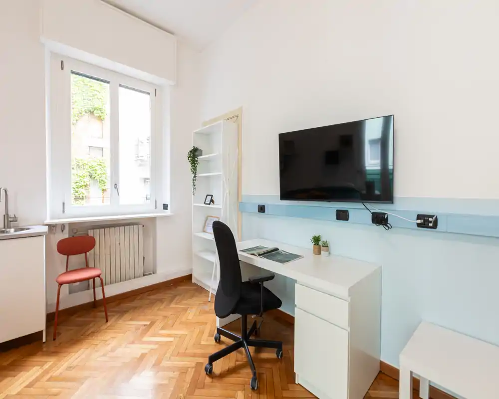 Spazio luminoso e moderno per lavoro/soggiorno con scrivania, TV a parete, scaffalatura e grande finestra che garantisce luce naturale — ideale per smart working o relax.
