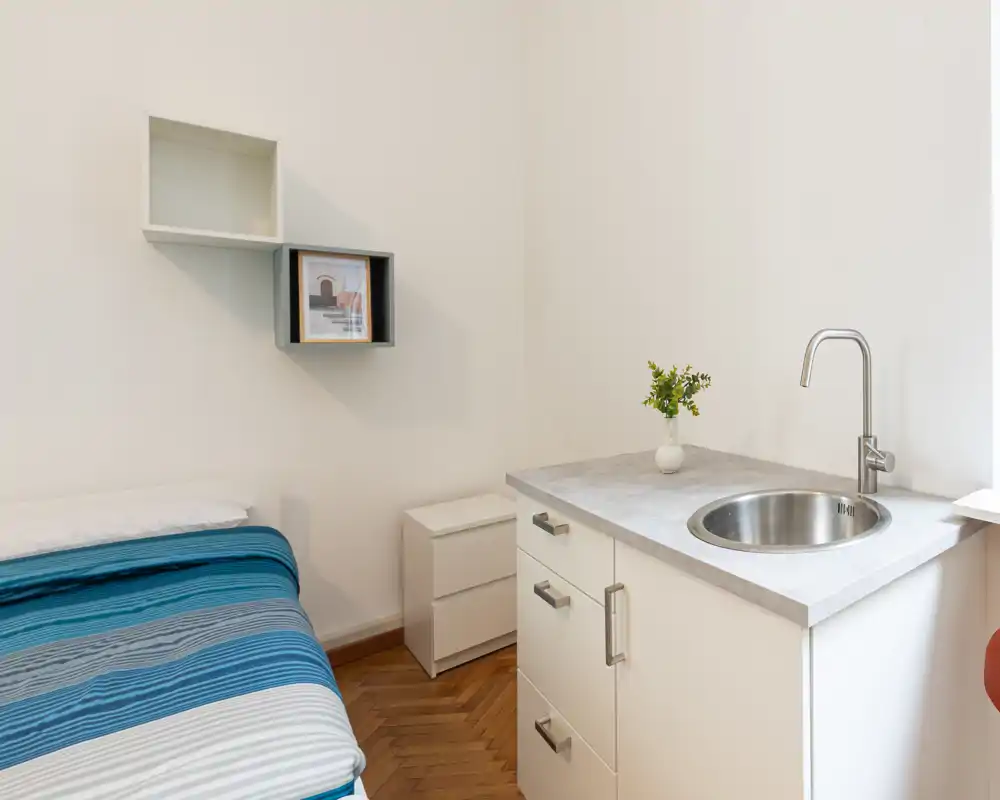 Camera compatta con letto singolo e angolo cucina dotato di lavabo rotondo e cassetti: spazio luminoso e arredato in modo semplice, ideale per un monolocale.