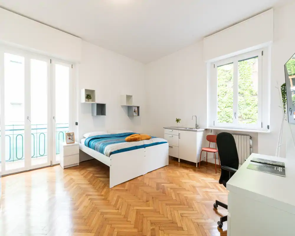 Camera da letto luminosa e spaziosa con letto matrimoniale, pavimento in parquet e grandi finestre che lasciano entrare molta luce naturale — arredi minimal e un piccolo angolo studio.