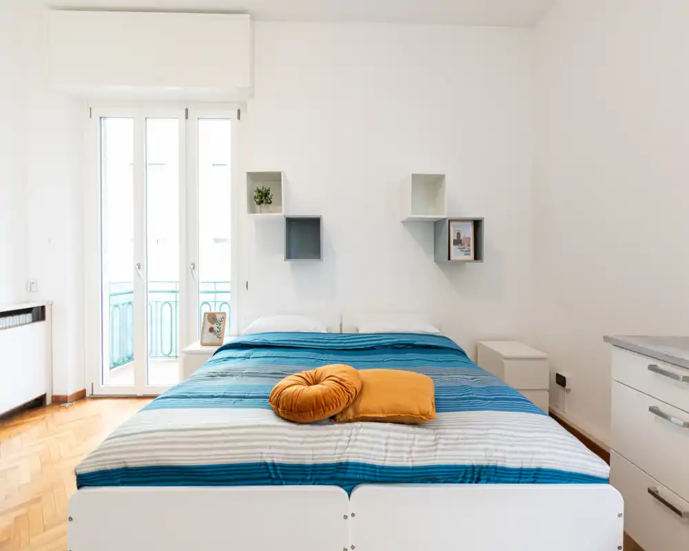 Camera da letto luminosa e moderna con un ampio letto matrimoniale con lenzuola a righe blu, cuscini decorativi color senape e luce naturale dalla porta del balcone. Mensole semplici e pareti bianche creano un'atmosfera pulita e rilassante, ideale per gli annunci.