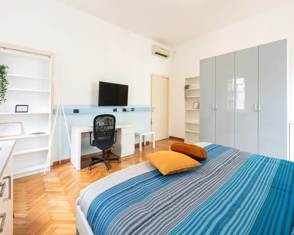 Camera da letto luminosa e moderna con ampio letto in primo piano, biancheria a righe colorata e cuscini decorativi, armadio e zona studio con TV e sedia — ideale per una o due persone in cerca di comfort e funzionalità.