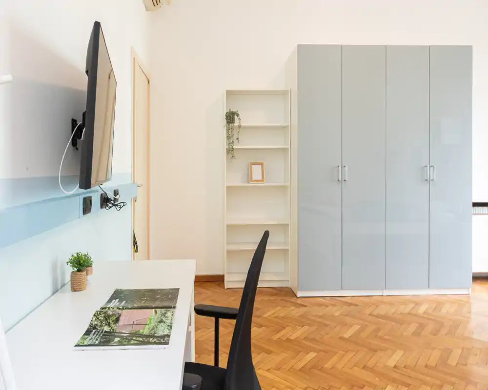 Angolo camera minimal con grande armadio, scaffale aperto e scrivania con sedia; pavimento in parquet luminoso e arredi moderni e semplici.