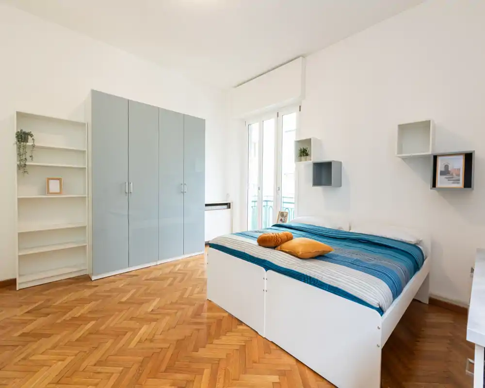 Camera da letto luminosa e moderna con letto matrimoniale con biancheria a righe blu, pavimento in parquet e un grande armadio; stile pulito e minimal con luce naturale dalla finestra-balcone.